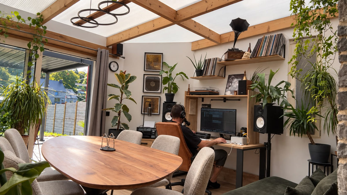 Adrien d'Elzius de dos, casque sur les oreilles, devant ses écrans et ses enceintes de studio, dans une véranda ouverte sur le jardin.
